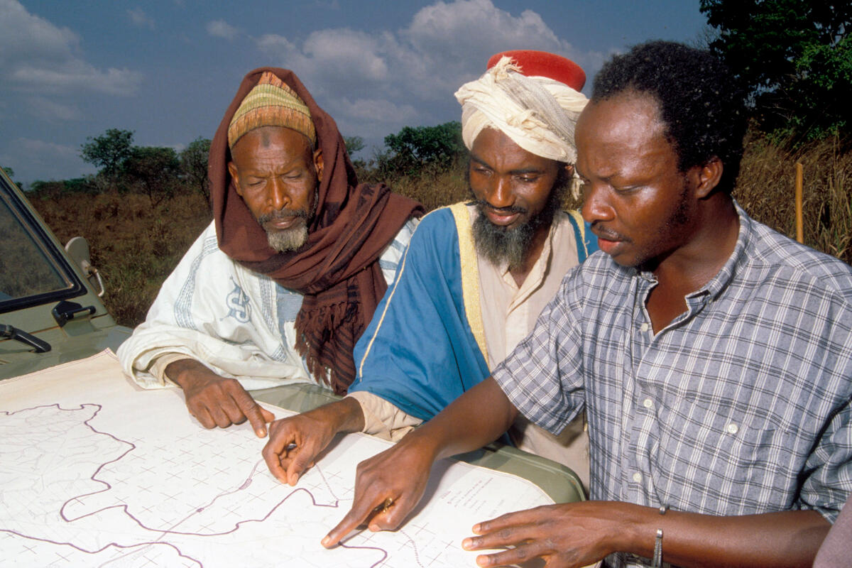 A man in city clothes helps two Central African herders trace grazing area boundaries on a map spread over a car's bonnet