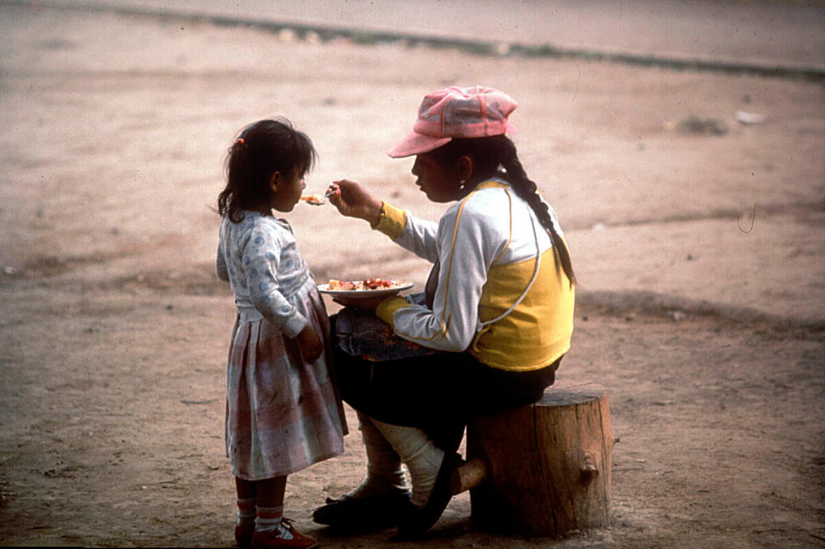 An Indigenous Ecuadorean woman sits on a tree stump feeding her young daughter from a plate