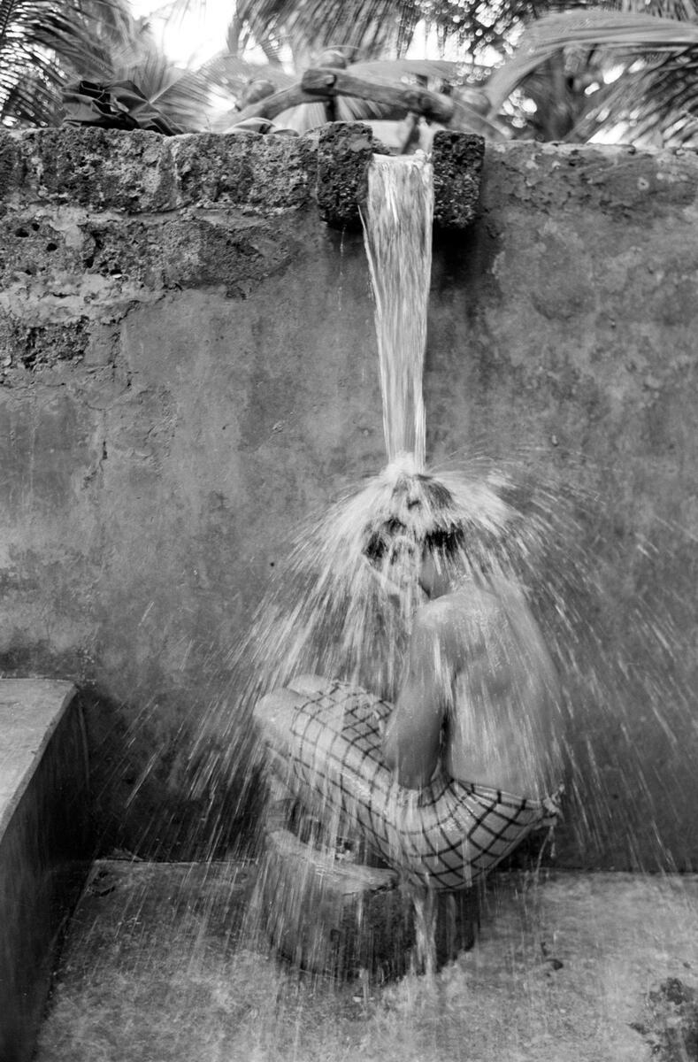 A rural Indian boy sits under a stream of water gushing from a pipe above him