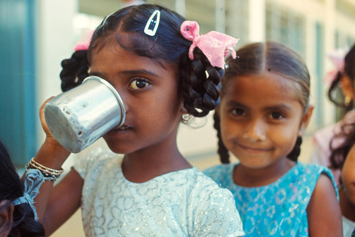 Two Mauritian primary school girls face the camera, one drinking from a tin mug