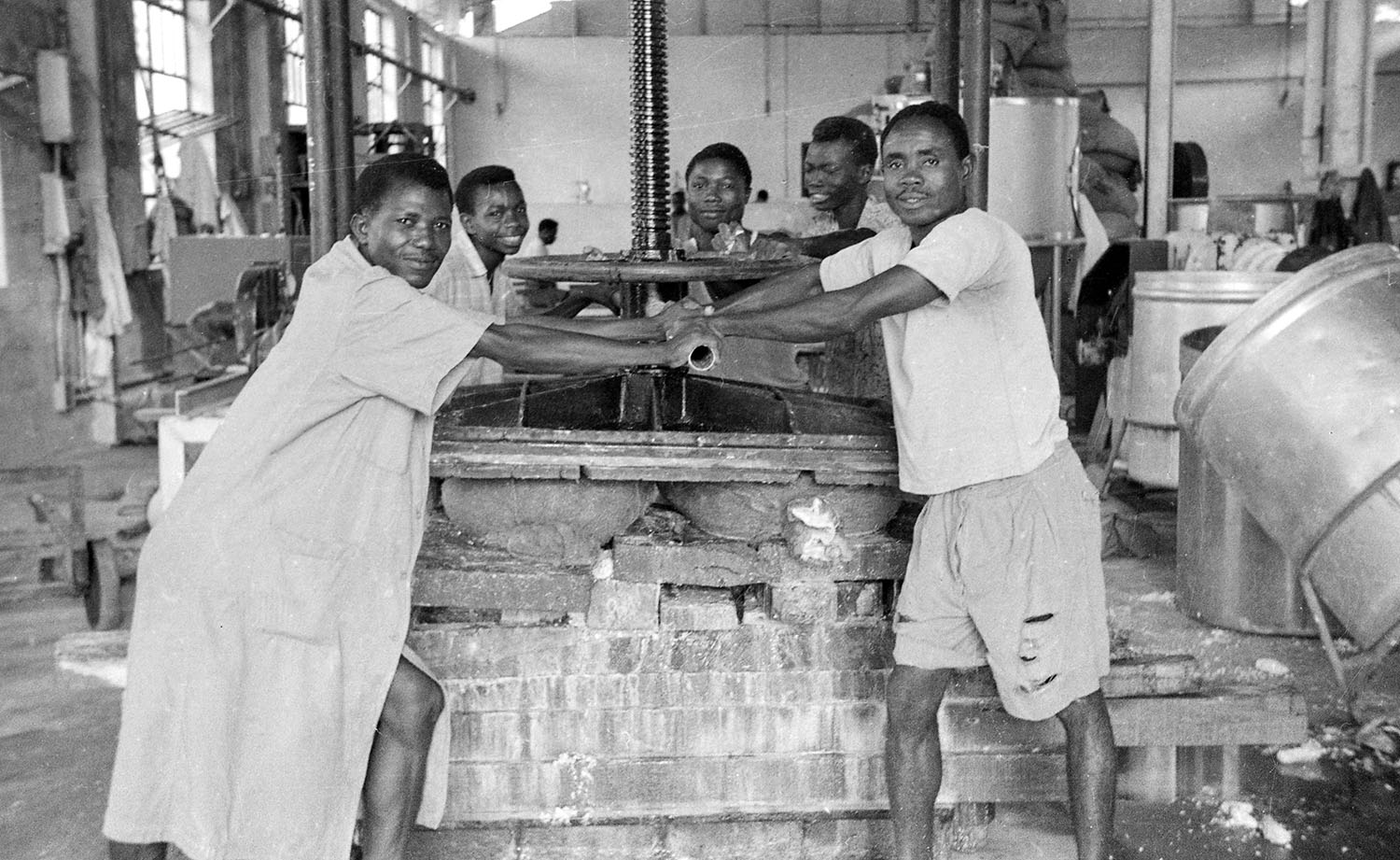 Five young Nigerian men smile at the camera as they operate a large manual cassava press